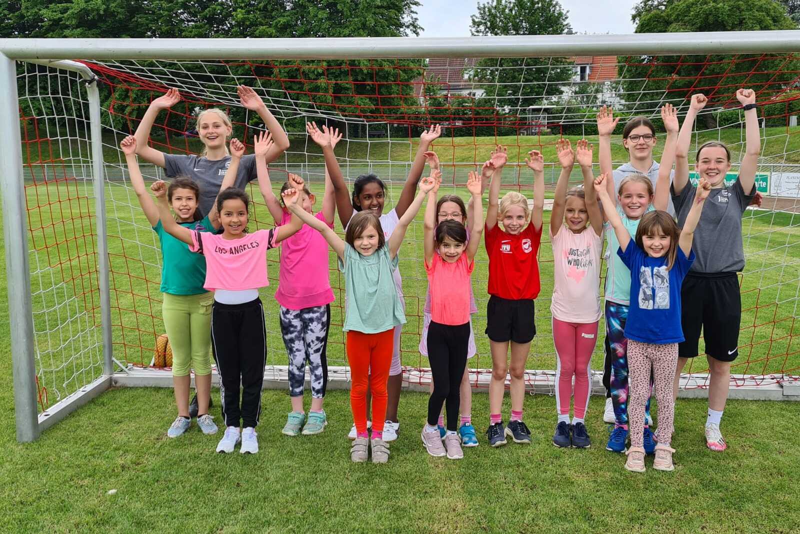 Gruppe von M&auml;dchen und Frauen feiert vor einem Fu&szlig;balltor, alle heben die Arme in die Luft.