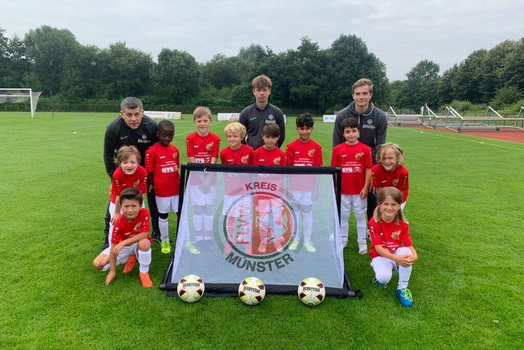 Gruppe von Fu&szlig;ballspielern in roten Trikots posiert vor einem Banner mit dem Schriftzug &bdquo;KREIS M&Uuml;NSTER&ldquo; auf einem Rasenplatz.