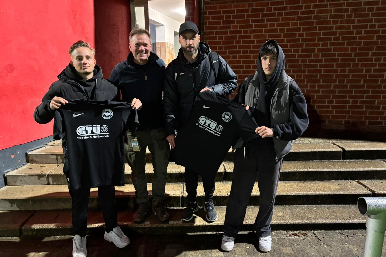 Vier Personen stehen auf einer Treppe vor einem roten Hintergrund und halten schwarze T-Shirts mit dem Aufdruck &bdquo;CTU&ldquo; in der Hand.