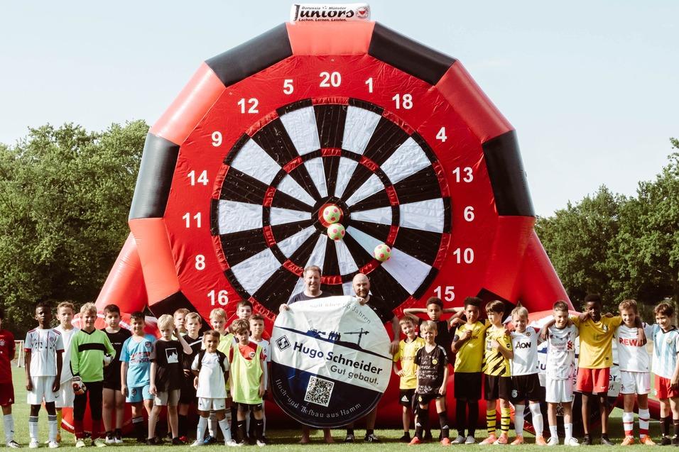 Gruppierung von Kindern im Fu&szlig;balloutfit vor einer gro&szlig;en, bunten Dartscheibe auf einem Sportplatz.