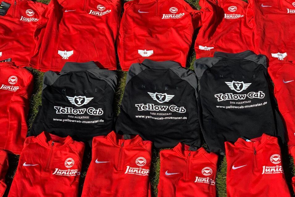 Eine Anordnung roter Trikots und schwarzer T-Shirts auf gr&uuml;nem Gras, einige mit Logos von Sponsoren.