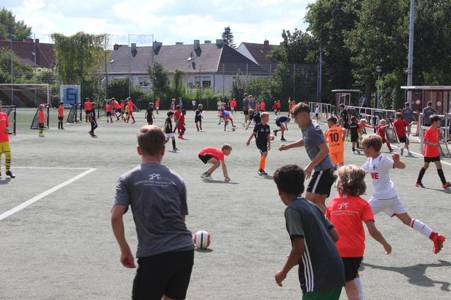 Gruppentraining von Kindern auf einem Fu&szlig;ballplatz, viele Spieler in bunten Shirts und Shorts, &uuml;ber verschiedene Aktivit&auml;ten verteilt.