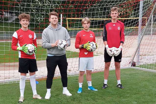 Drei Fu&szlig;ballspieler in roten Trikots stehen mit B&auml;llen vor einem Fu&szlig;balltor auf einem Rasenplatz.