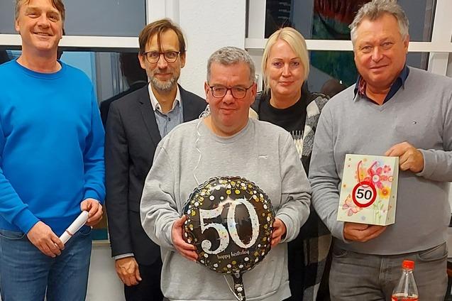 F&uuml;nf Personen stehen zusammen, eine Person h&auml;lt einen schwarzen Luftballon mit der Zahl 50, im Hintergrund sind Geschenke.