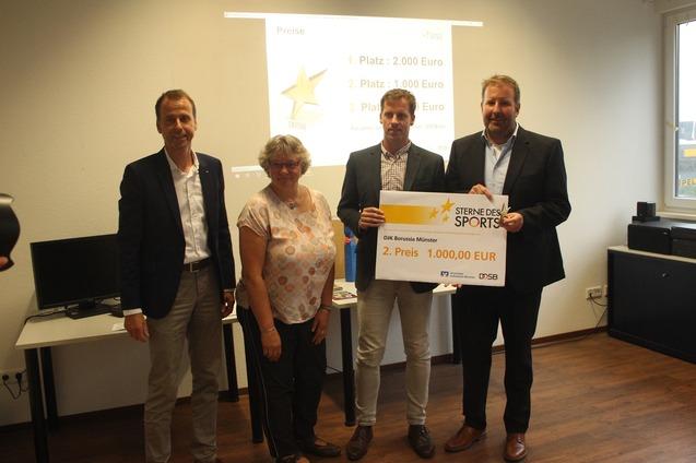 Vier Personen stehen in einem Raum und halten ein gro&szlig;es Preis-Zertifikat f&uuml;r den zweiten Platz beim Wettbewerb &bdquo;Sterne des Sports&ldquo;.