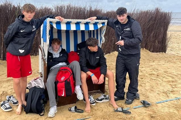 Vier Jugendliche stehen am Strand, einer sitzt in einem Strandkorb, alle tragen sportliche Kleidung und Flip-Flops.