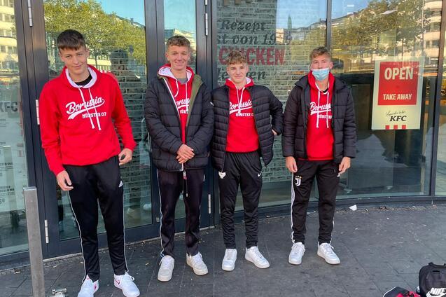 Vier Jugendliche in roten Sweatshirts und Sporthosen stehen vor einem Restaurant mit gro&szlig;en Fenstern.