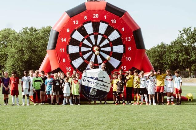 Gruppierung von Kindern im Fu&szlig;balloutfit vor einer gro&szlig;en, bunten Dartscheibe auf einem Sportplatz.