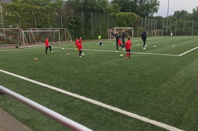 Gruppe von Kindern in roten Trainingsanz&uuml;gen &uuml;bt mit Fu&szlig;b&auml;llen auf einem Fu&szlig;ballfeld mit Kunstrasen.