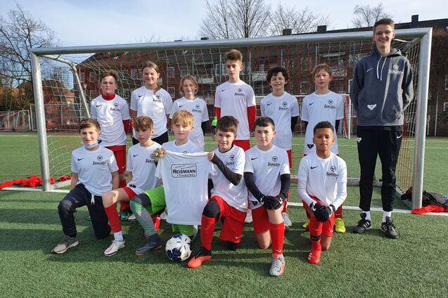 Gruppe von zw&ouml;lf Jugendlichen in wei&szlig;en Shirts mit roten Hosen, posierend vor einem Fu&szlig;balltor auf einem Platz.