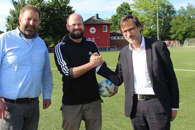 Drei M&auml;nner sch&uuml;tteln sich die H&auml;nde auf einem Fu&szlig;ballplatz, im Hintergrund ein Sportgeb&auml;ude und B&auml;ume.