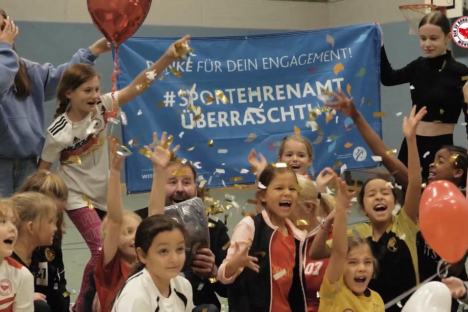 Gruppenfoto von l&auml;chelnden Kindern und Jugendarbeitern, umgeben von Luftballons und Konfetti, vor einem Banner &uuml;ber Ehrenamt.
