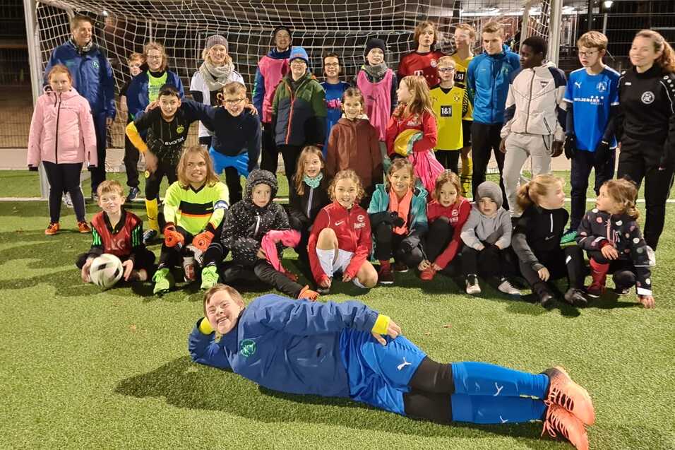 Gruppenfoto von Kindern und Trainern auf einem Fu&szlig;ballplatz, alle tragen Sportkleidung und stehen vor dem Tor.