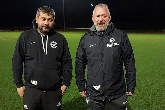Zwei Trainer stehen auf einem beleuchteten, gr&uuml;nen Fu&szlig;ballfeld in der Dunkelheit.