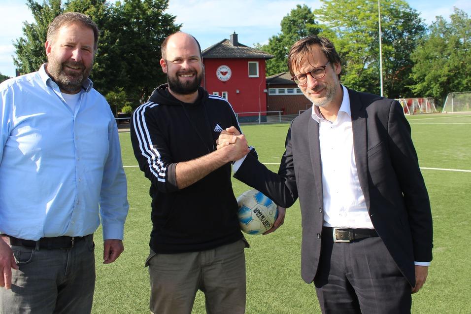 Drei M&auml;nner sch&uuml;tteln sich die H&auml;nde auf einem Fu&szlig;ballplatz, im Hintergrund ein Sportgeb&auml;ude und B&auml;ume.