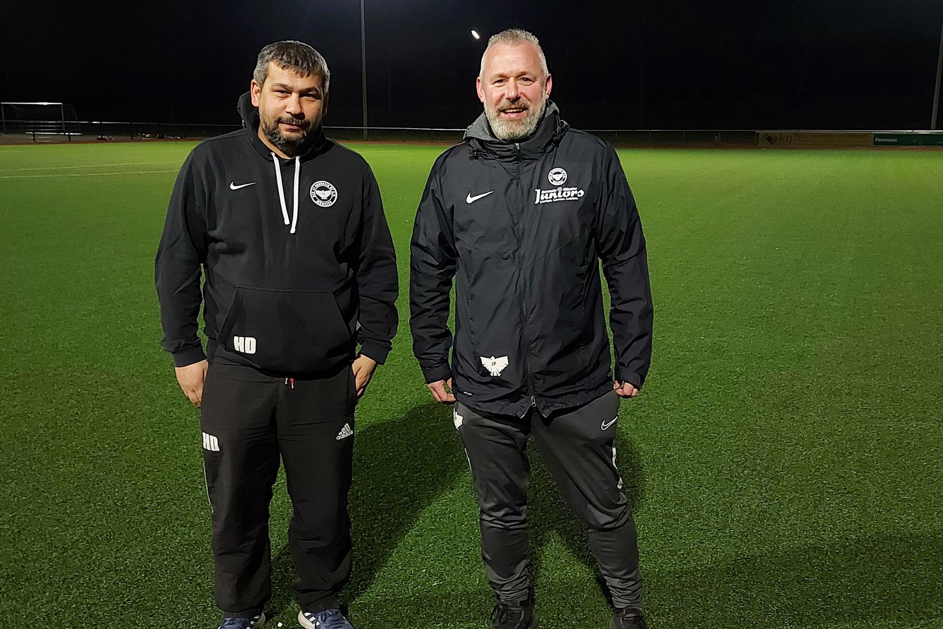 Zwei Trainer stehen auf einem beleuchteten, gr&uuml;nen Fu&szlig;ballfeld in der Dunkelheit.