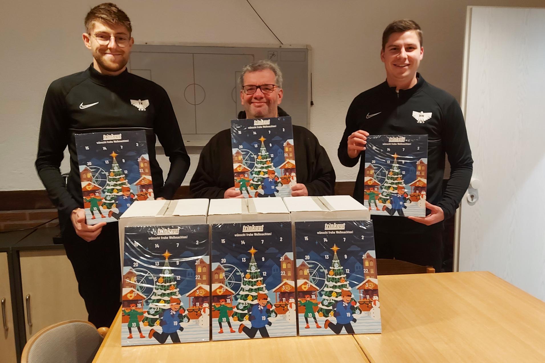 Drei M&auml;nner in schwarz tragen Adventskalender vor einer Tischplatte mit Weihnachtsmotiv und stehen in einem Raum.