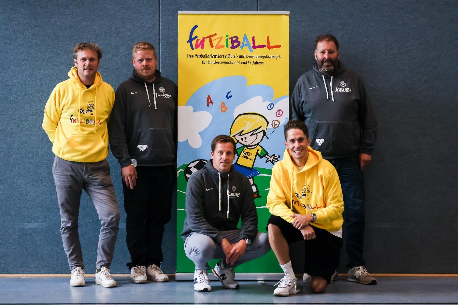 F&uuml;nf M&auml;nner in Sportswear stehen vor einem Banner mit der Aufschrift &bdquo;FurtziBALL&ldquo; und einem Kindermotiv.