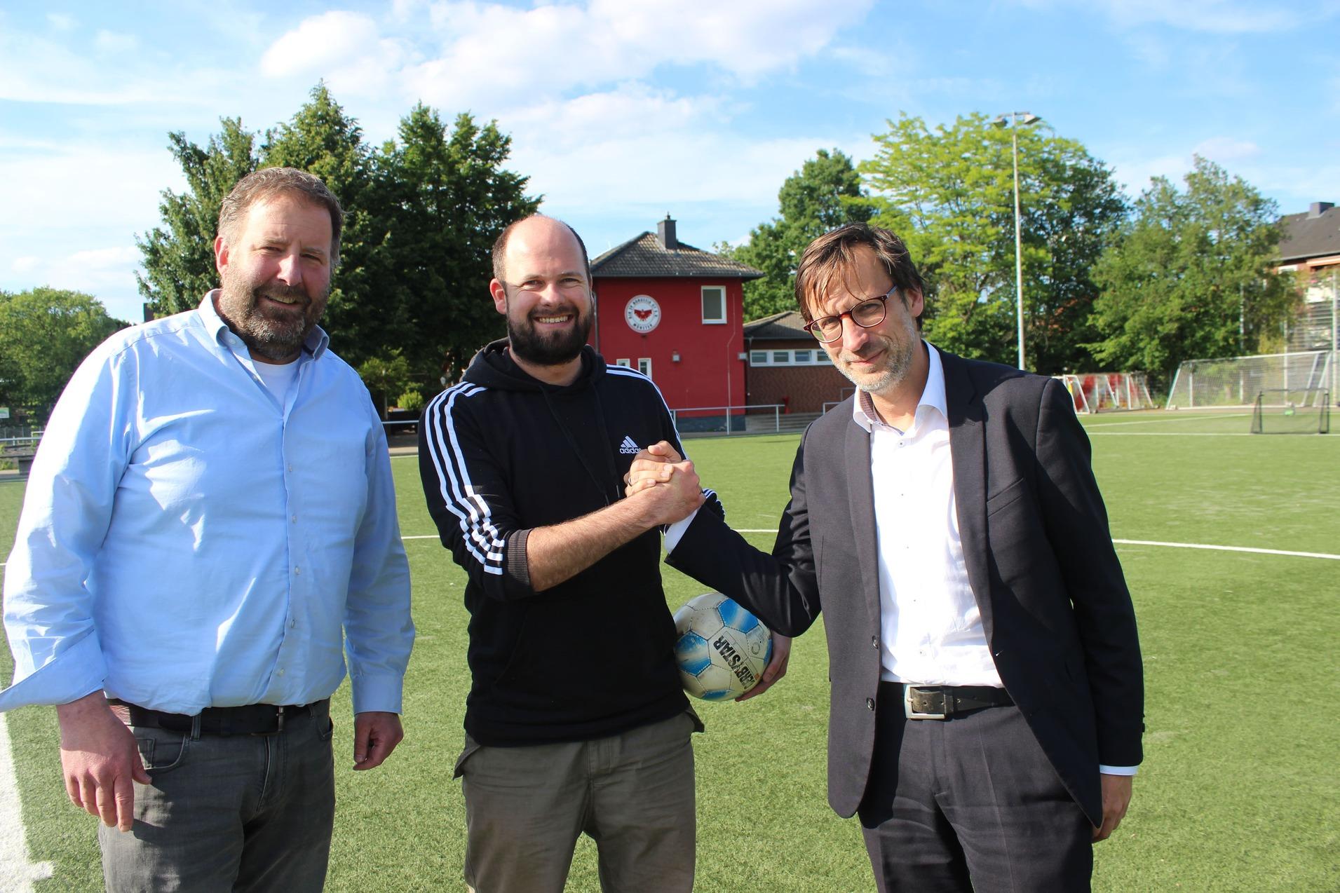 Drei M&auml;nner sch&uuml;tteln sich die H&auml;nde auf einem Fu&szlig;ballplatz, im Hintergrund ein Sportgeb&auml;ude und B&auml;ume.