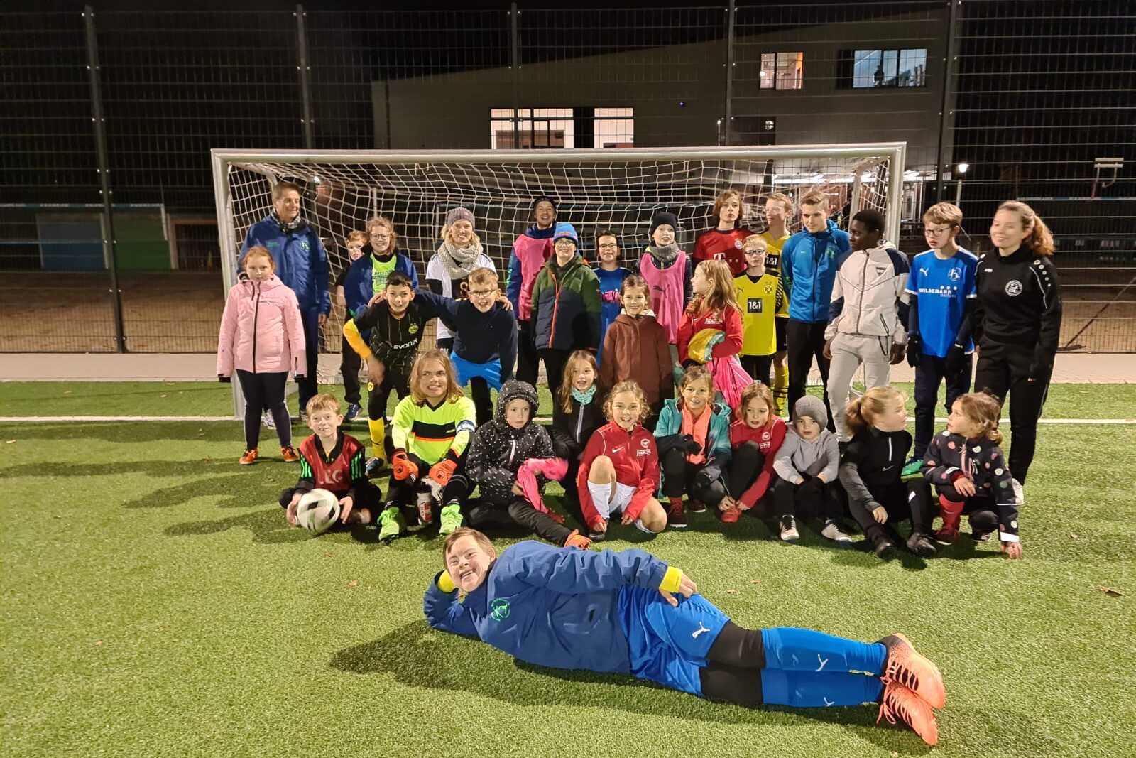 Gruppenfoto von Kindern und Trainern auf einem Fu&szlig;ballplatz, alle tragen Sportkleidung und stehen vor dem Tor.