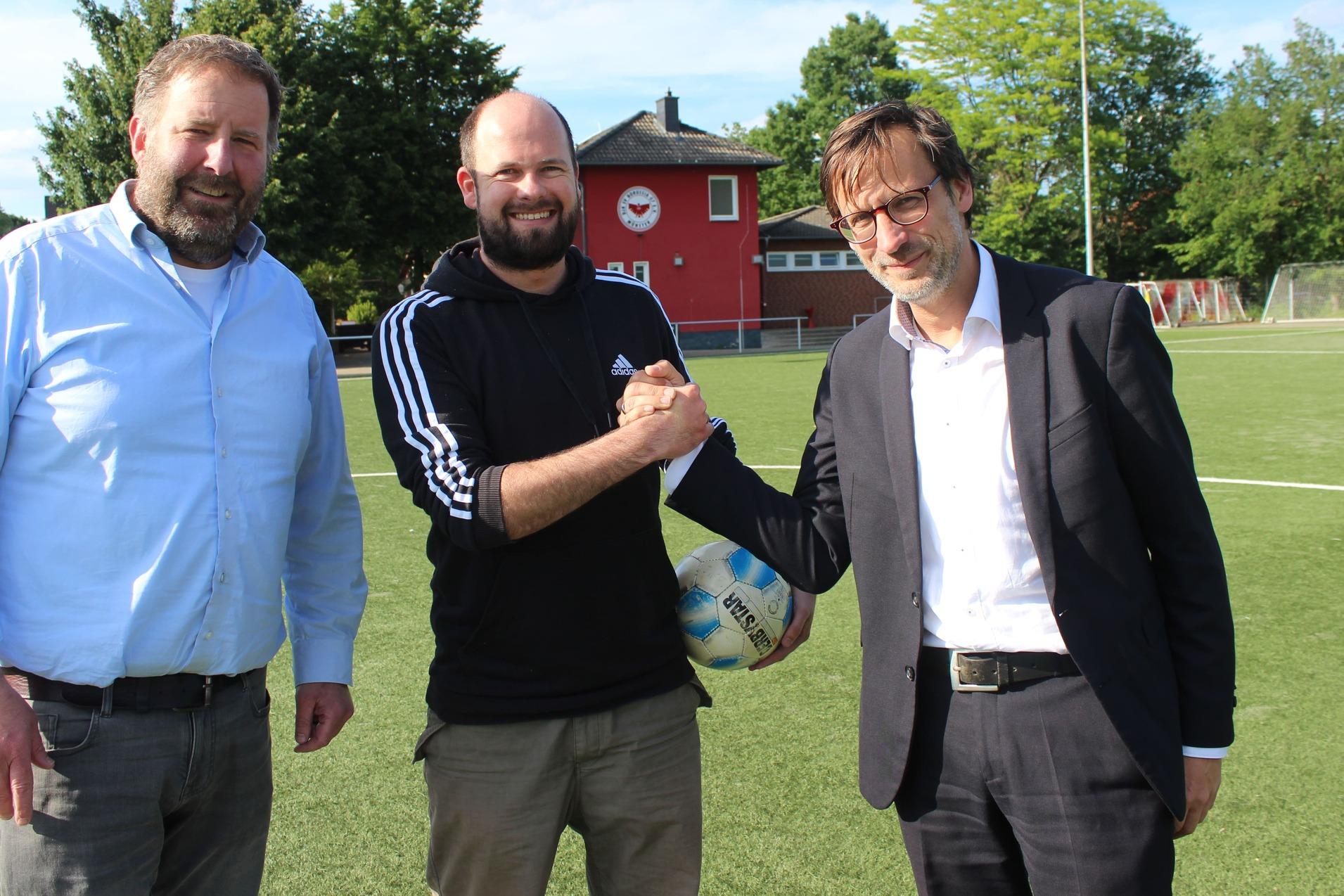 Drei Männer schütteln sich die Hände auf einem Fußballplatz, im Hintergrund ein Sportgebäude und Bäume.