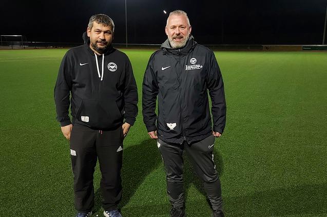 Zwei Trainer stehen auf einem beleuchteten, gr&uuml;nen Fu&szlig;ballfeld in der Dunkelheit.