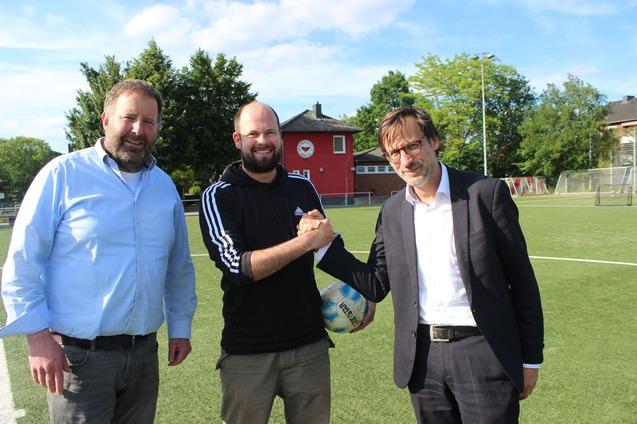 Drei M&auml;nner sch&uuml;tteln sich die H&auml;nde auf einem Fu&szlig;ballplatz, im Hintergrund ein Sportgeb&auml;ude und B&auml;ume.