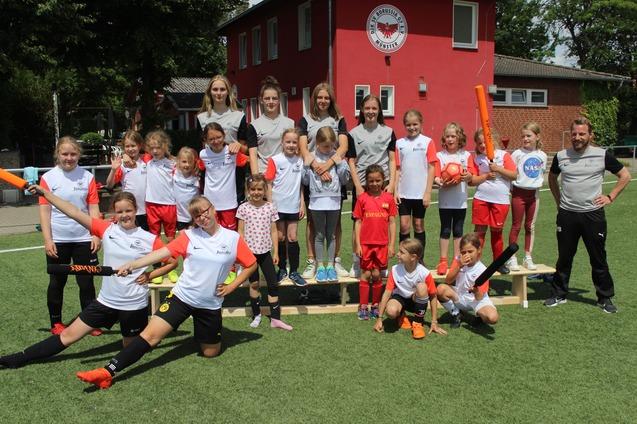 Gruppe von Fu&szlig;ballspielerinnen und -spielern auf einem Sportplatz, einige halten orangefarbene Sportger&auml;te.