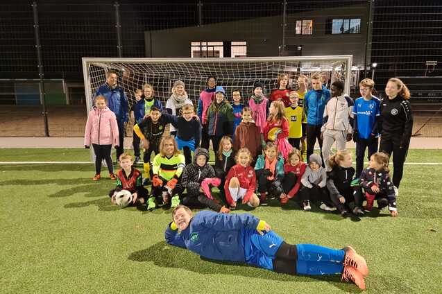 Gruppenfoto von Kindern und Trainern auf einem Fu&szlig;ballplatz, alle tragen Sportkleidung und stehen vor dem Tor.