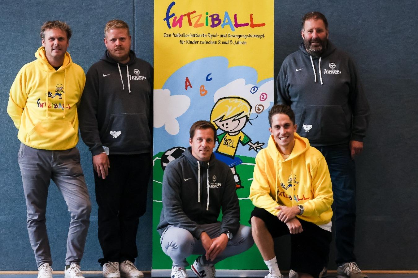 Fünf Männer in Sportswear stehen vor einem Banner mit der Aufschrift „FurtziBALL“ und einem Kindermotiv.