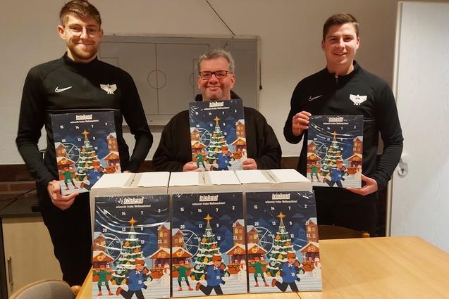 Drei M&auml;nner in schwarz tragen Adventskalender vor einer Tischplatte mit Weihnachtsmotiv und stehen in einem Raum.