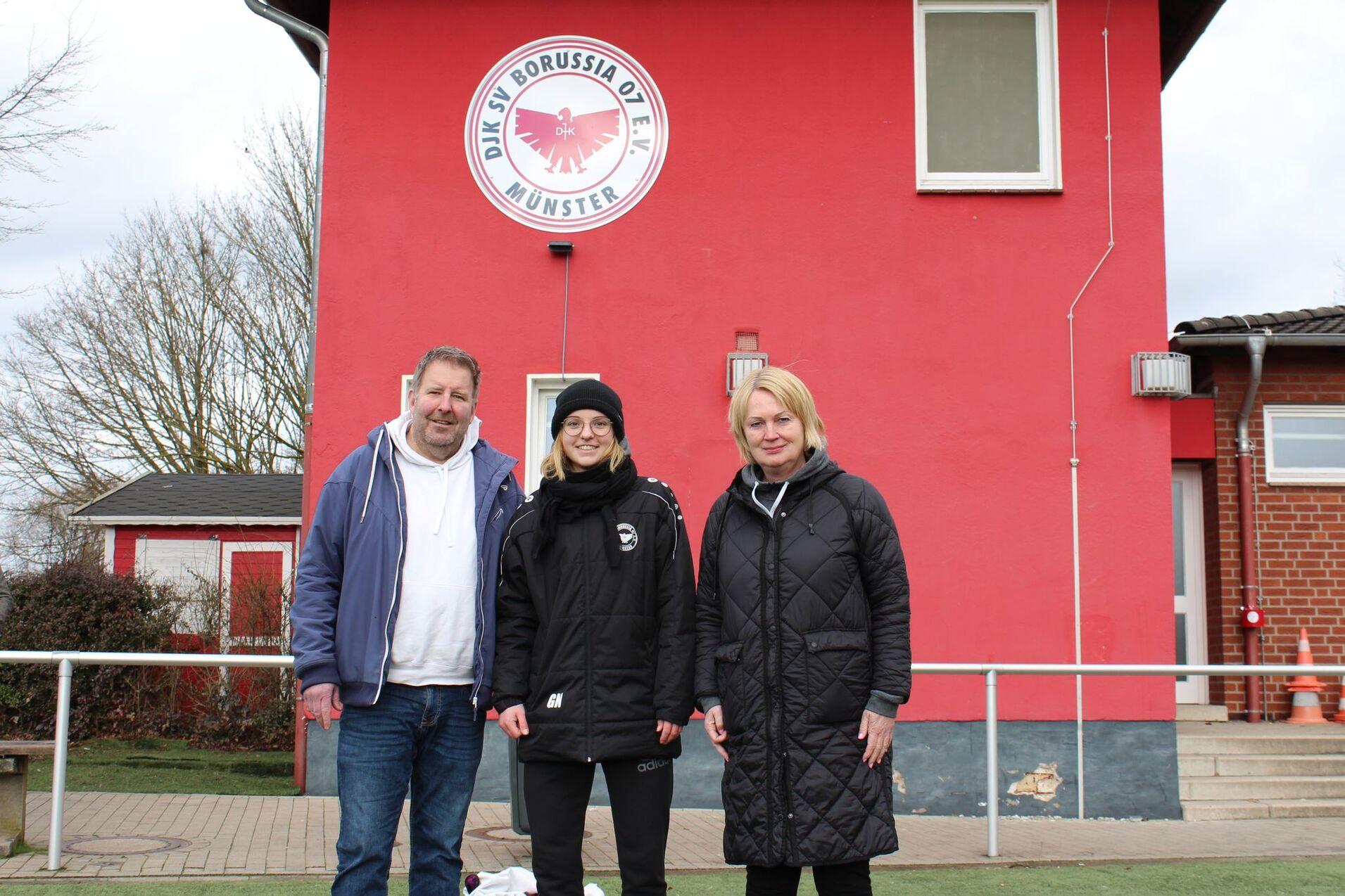 Drei Personen stehen vor einem roten Vereinsgeb&auml;ude mit dem Logo von Borussia M&uuml;nster.