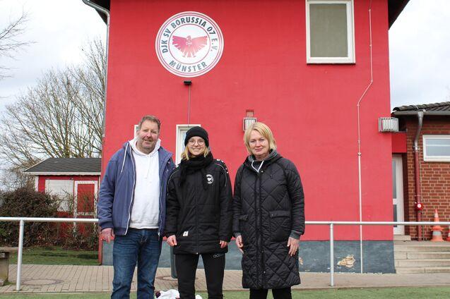 Drei Personen stehen vor einem roten Vereinsgeb&auml;ude mit dem Logo von Borussia M&uuml;nster.