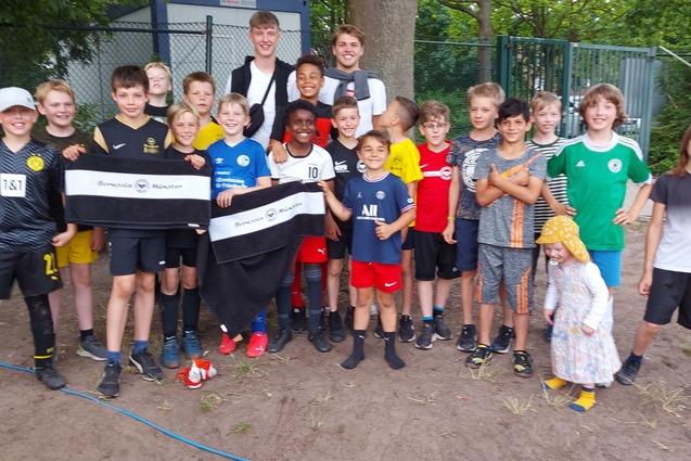 Gruppe von 18 Jungen in Fu&szlig;ballkleidung posiert zusammen im Freien, einige halten Banner.
