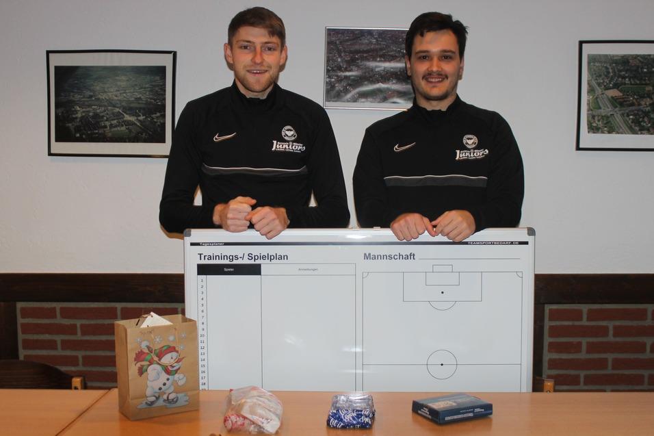 Zwei Fu&szlig;balltrainer stehen hinter einem gro&szlig;en Trainingsspielplan, w&auml;hrend Geschenke und S&uuml;&szlig;igkeiten auf dem Tisch liegen.