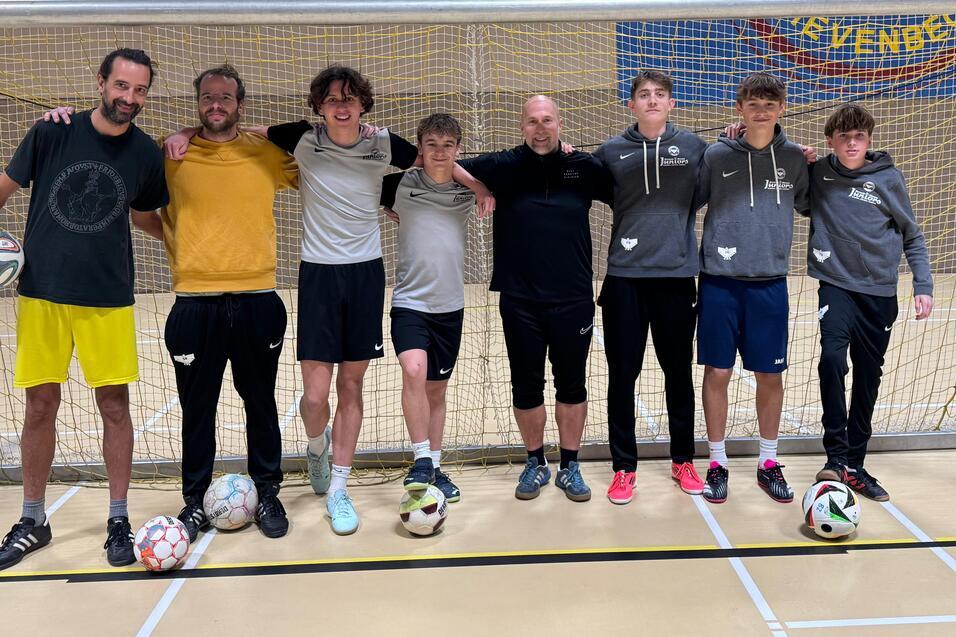 Gruppe von acht Personen in Sportkleidung steht l&auml;chelnd vor einem Fu&szlig;balltor in einer Halle, B&auml;lle zu ihren F&uuml;&szlig;en.