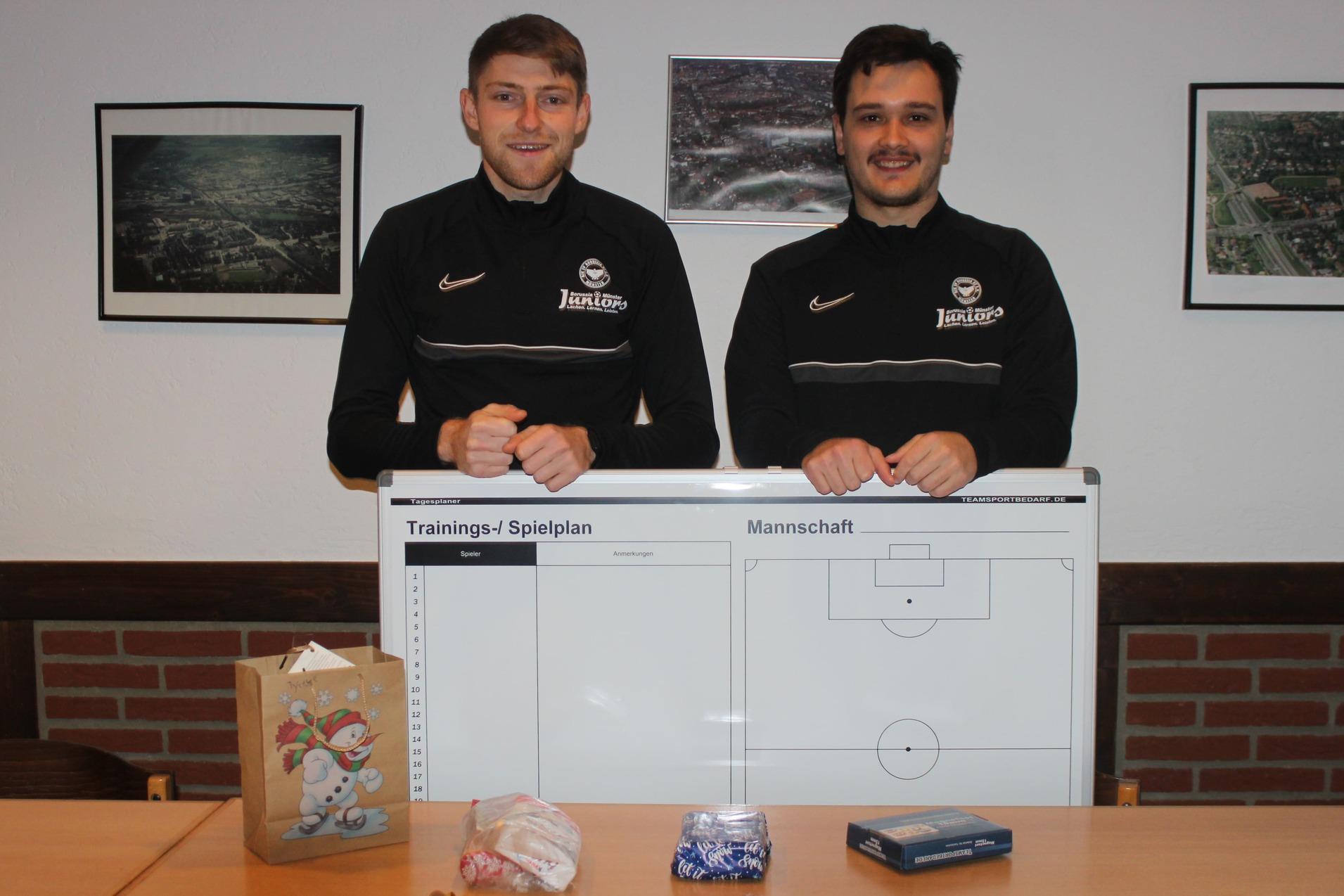 Zwei Fu&szlig;balltrainer stehen hinter einem gro&szlig;en Trainingsspielplan, w&auml;hrend Geschenke und S&uuml;&szlig;igkeiten auf dem Tisch liegen.