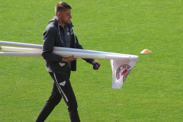 Mann tr&auml;gt mehrere wei&szlig;e Rohre und eine Flagge &uuml;ber den gr&uuml;nen Fu&szlig;ballplatz.