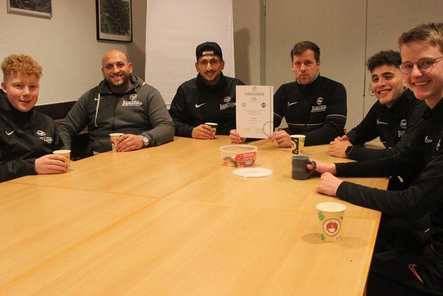 Sechs Personen in Sportkleidung sitzen an einem Tisch, halten Getr&auml;nke und blicken auf ein Dokument.