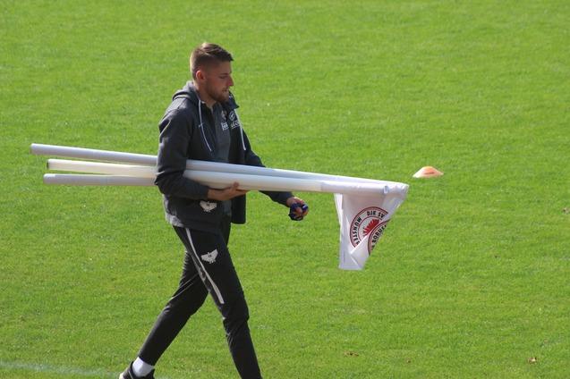 Mann tr&auml;gt mehrere wei&szlig;e Rohre und eine Flagge &uuml;ber den gr&uuml;nen Fu&szlig;ballplatz.