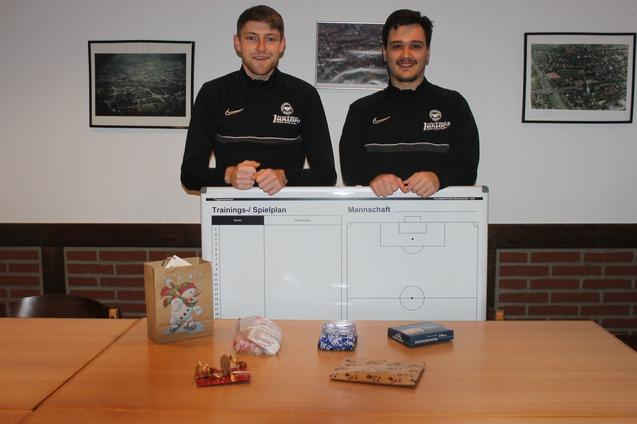 Zwei Fu&szlig;balltrainer stehen hinter einem gro&szlig;en Trainingsspielplan, w&auml;hrend Geschenke und S&uuml;&szlig;igkeiten auf dem Tisch liegen.