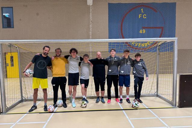Gruppe von acht Personen in Sportkleidung steht l&auml;chelnd vor einem Fu&szlig;balltor in einer Halle, B&auml;lle zu ihren F&uuml;&szlig;en.