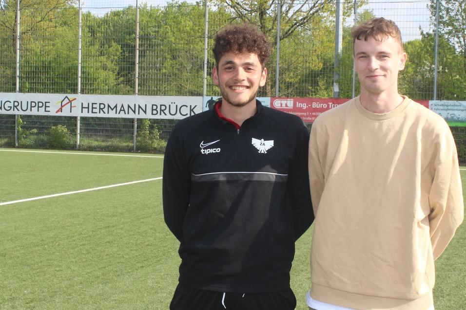 Zwei junge M&auml;nner stehen l&auml;chelnd auf einem Fu&szlig;ballfeld, im Hintergrund sind Sponsorenbanner und B&auml;ume zu sehen.