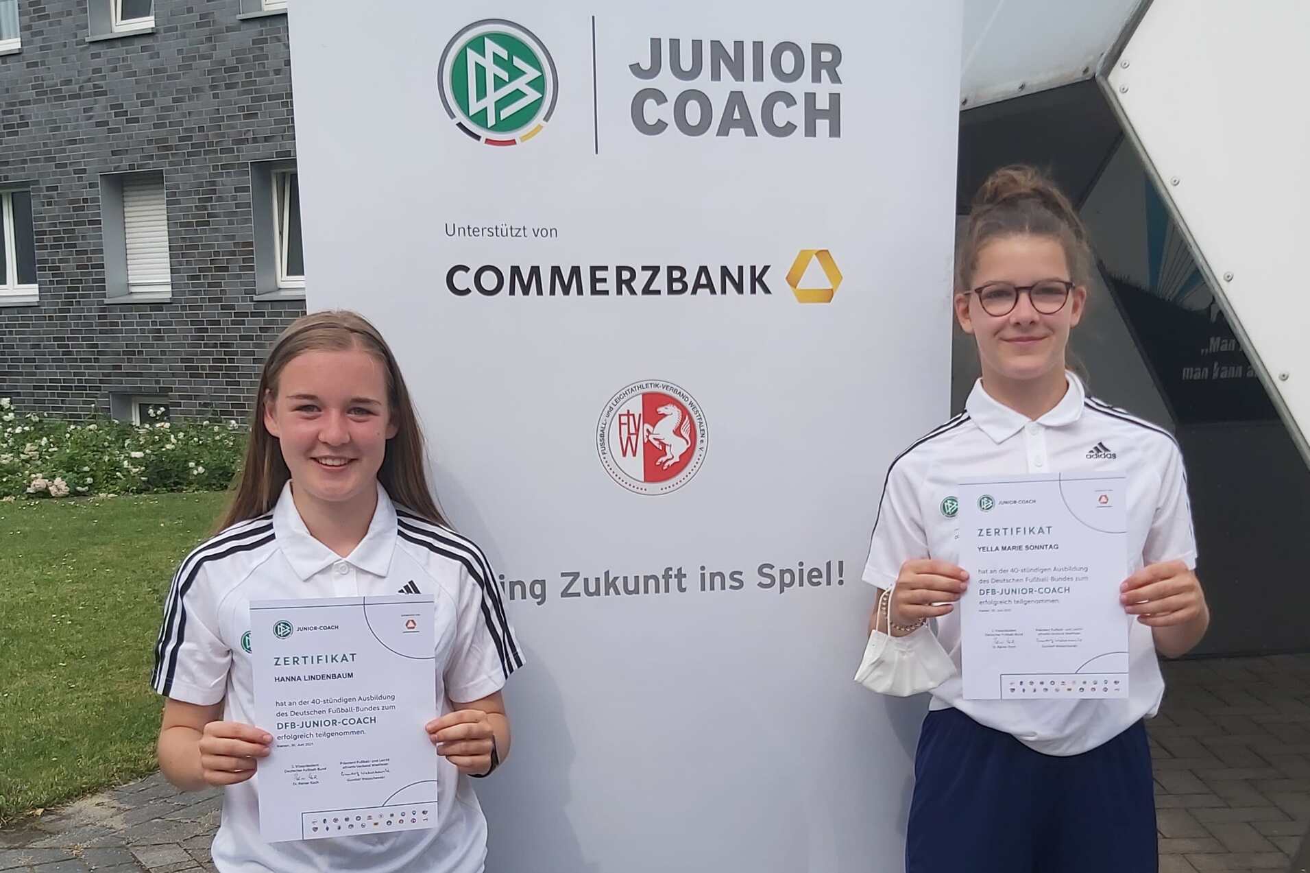 Zwei junge Fu&szlig;ballerinnen halten stolz ihre Bescheinigungen vor einem Banner der Junior Coach-Initative.