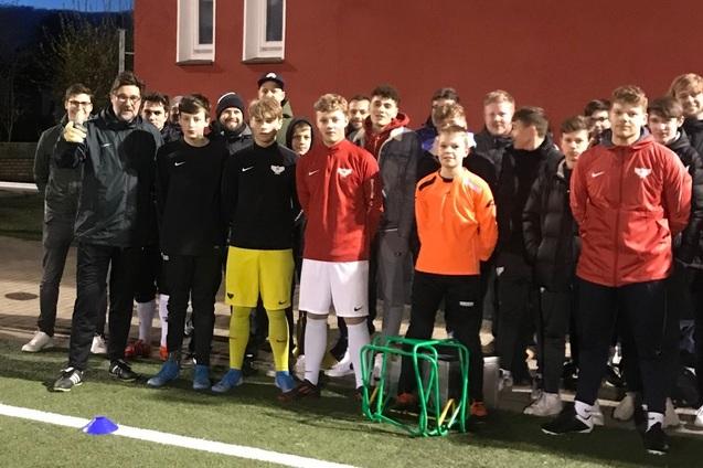 Gruppe von Fu&szlig;ballspielern in Trainingskleidung posiert vor einer Sportanlage bei D&auml;mmerung.