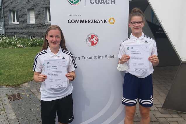 Zwei junge Fu&szlig;ballerinnen halten stolz ihre Bescheinigungen vor einem Banner der Junior Coach-Initative.