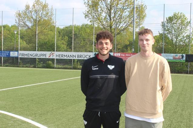 Zwei junge M&auml;nner stehen l&auml;chelnd auf einem Fu&szlig;ballfeld, im Hintergrund sind Sponsorenbanner und B&auml;ume zu sehen.
