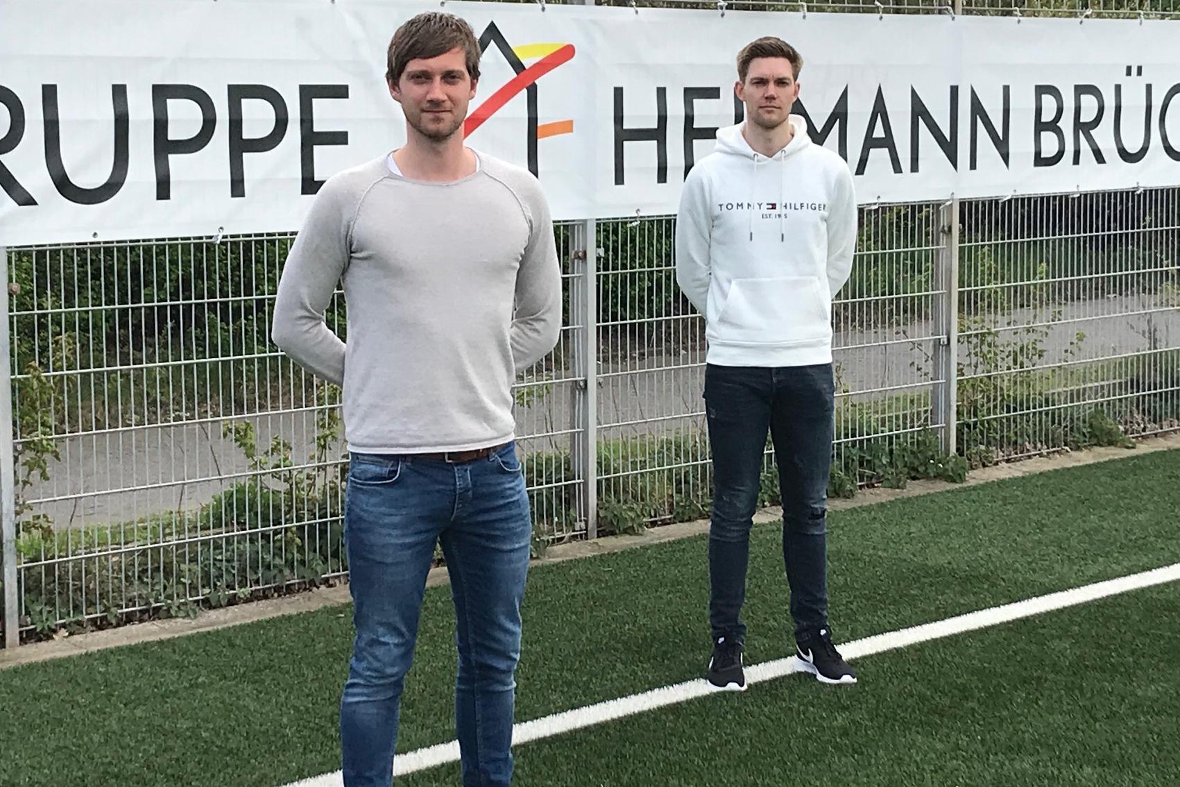 Zwei junge M&auml;nner stehen auf einem Kunstrasenfeld vor einem Banner mit der Aufschrift &bdquo;GRUPPE HERMANN BR&Uuml;CK&ldquo;.