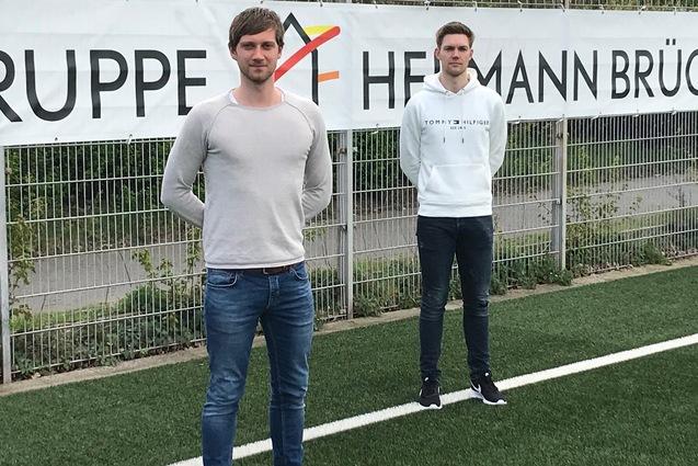 Zwei junge M&auml;nner stehen auf einem Kunstrasenfeld vor einem Banner mit der Aufschrift &bdquo;GRUPPE HERMANN BR&Uuml;CK&ldquo;.