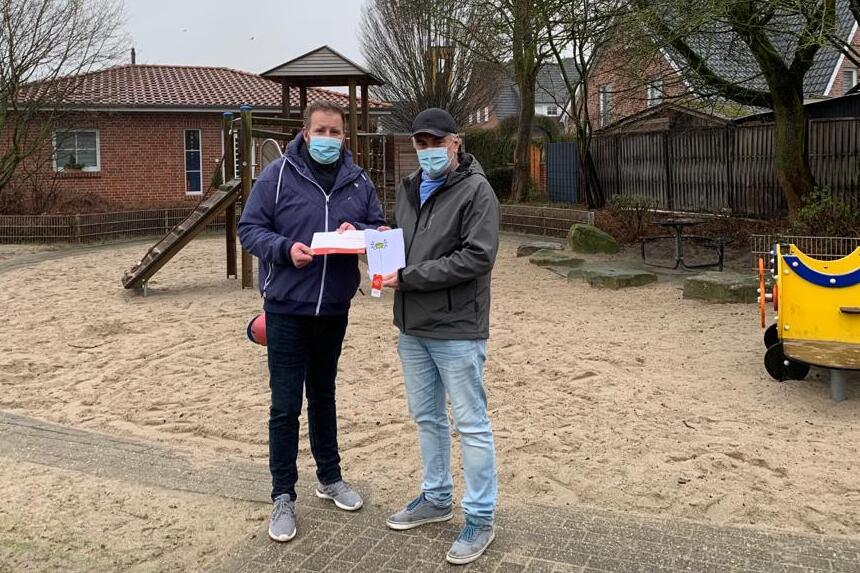 Zwei Personen mit Mund-Nasen-Schutz stehen auf einem Sandspielplatz und halten ein Dokument in der Hand.